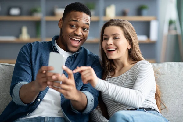 Nadšený multikulturní pár pomocí smartphonu společně při relaxaci na gauči doma — Stock fotografie