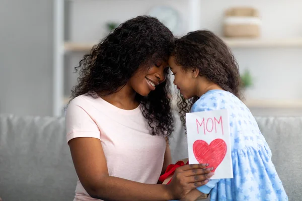 Zärtliches schwarzes Mädchen, das seiner Mutter zu Hause ein verpacktes Geburtstagsgeschenk und eine Grußkarte schenkt. Festliche Familientradition — Stockfoto