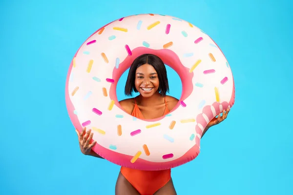 Young black lady in swimsuit posing with donut shaped inflatable ring, smiling at camera over blue studio background — Stock Photo, Image