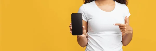 Neue mobile App. Schwarze Dame zeigt auf Smartphone mit leerem Bildschirm auf gelbem Hintergrund, Panorama mit freiem Platz — Stockfoto