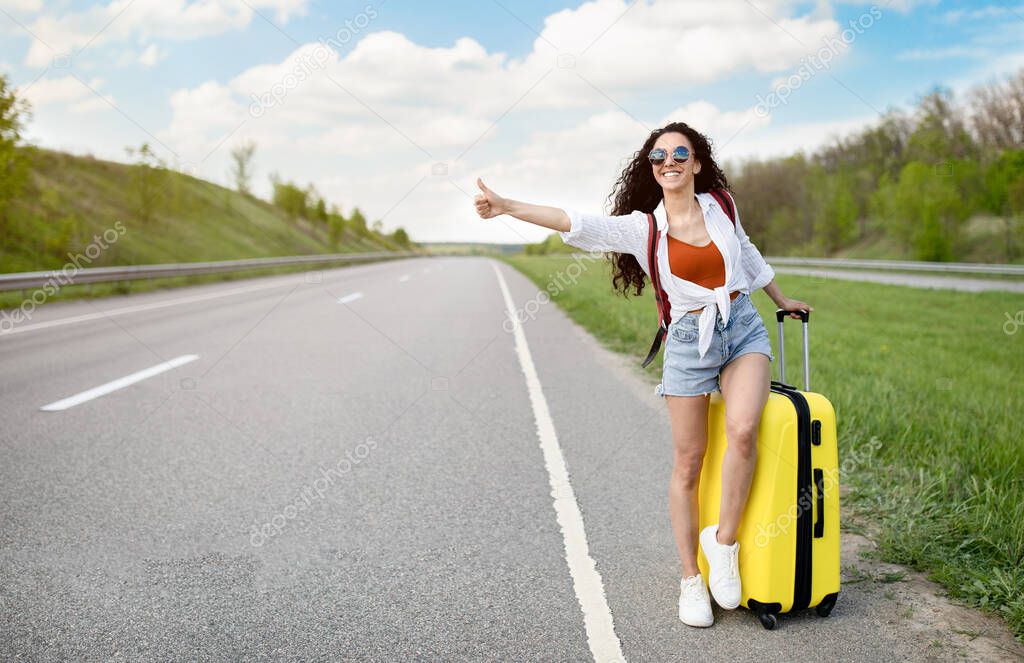Happy millennial woman with backpack and suitcase trying to stop passing car, needing free ride, hitchhiking on roadside
