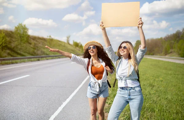 Két női barát potyautaskodik, üres kartontáblával, álarccal, az út szélén álló autóval, másolja a helyet. — Stock Fotó
