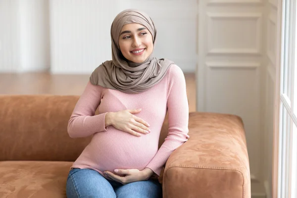 Vidám terhes muszlim nő, Hidzsábot visel, megérinti a hasát otthon — Stock Fotó
