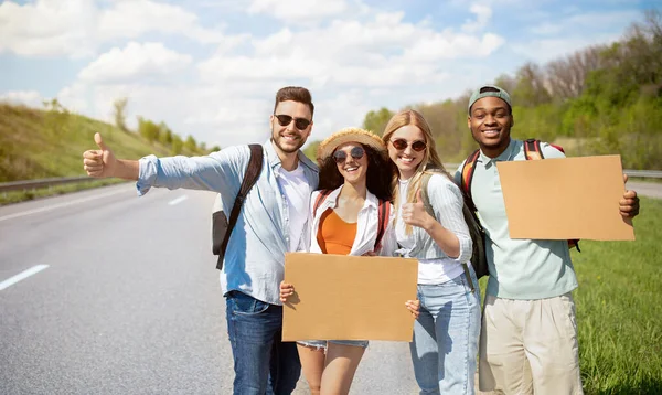 Retrato de vários amigos felizes carona juntos na estrada, fazendo gestos polegar para cima, segurando sinais vazios — Fotografia de Stock