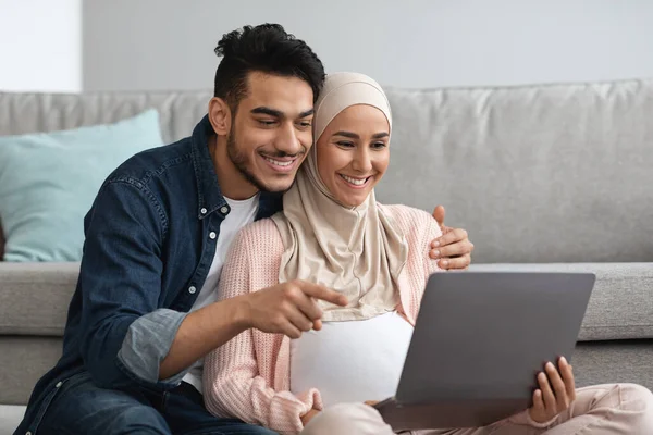 Веселая беременная мусульманка и ее муж пользуются ноутбуком дома вместе — стоковое фото