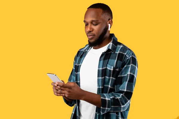 Black Millennial Male Menggunakan Cellphone Memakai Earbuds On Yellow Background — Stok Foto