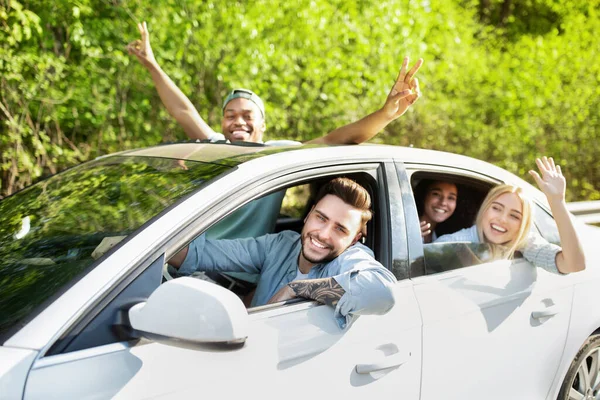 Fröhliche junge Freunde fahren Auto, blicken durch Fenster, winken und gestikulieren, gehen auf Roadtrip — Stockfoto