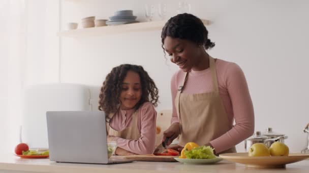 Mujer afroamericana feliz cortando verduras en la cocina con su hija pequeña, proceso de transmisión en línea a través de la computadora portátil — Vídeo de stock