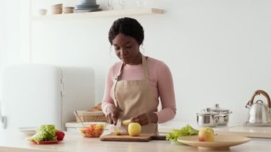 Önlük takan, elma yiyen, organik atıştırmalıkları seven, mutfakta dans eden Afro-Amerikan genç bayan.