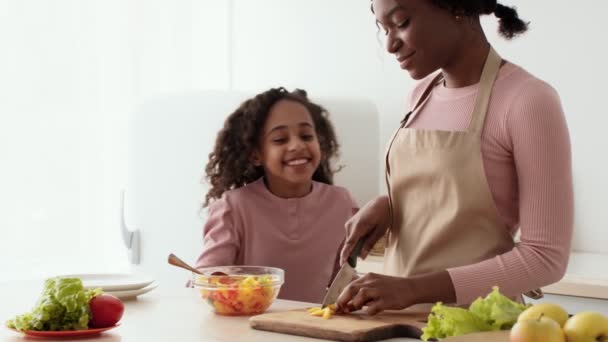 Mladá africká americká matka připravuje zeleninový salát, kudrnatá dcerka přichází a jí čerstvý pepř v kuchyni — Stock video
