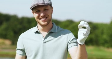 Profesyonel eldivenler giymiş, golf topu tutan ve golf sahasında kameraya gülümseyen beyaz şapkalı genç bir adam.