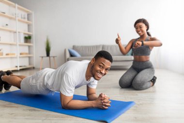 Sportif siyahi adam tahta duruşta kameraya gülümsüyor, kız arkadaşı onu akıllı saatle izliyor, başparmağını gösteriyor.