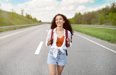 Yazlık giysiler içinde neşeli genç bir bayan kırsalda asfalt yolda yürüyor, dışarıda otostop çekiyor.