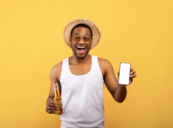 Funky cara preto em roupas de verão segurando celular com tela em branco e garrafa de cerveja no fundo amarelo, mockup — Fotografia de Stock