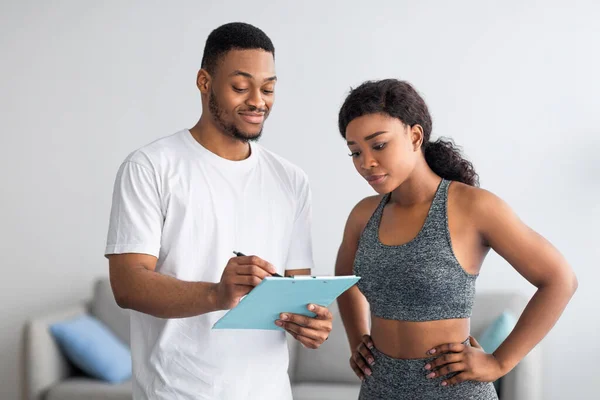 Schwarzer Personal Trainer füllt Trainingsplaner für junge Dame zu Hause aus — Stockfoto