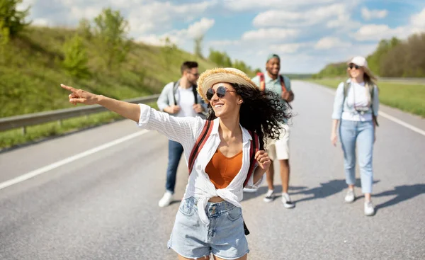 Mulher caucasiana jovem com seus amigos multirraciais carona na estrada, viajando por autostop, ao ar livre — Fotografia de Stock
