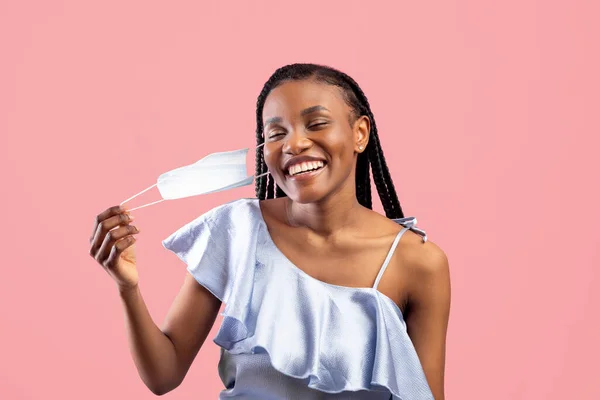 El coronavirus ha terminado. Mujer negra feliz con racimos afro quitándose la mascarilla en el fondo rosa del estudio — Foto de Stock