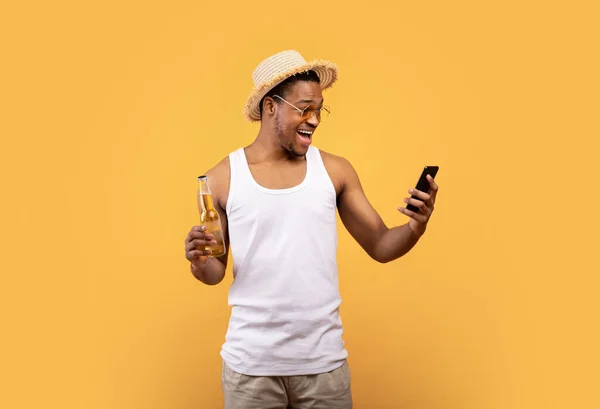 Jovem negro com garrafa de cerveja olhando para o celular em emoção, loteria vencedora ou cassino, fundo amarelo — Fotografia de Stock