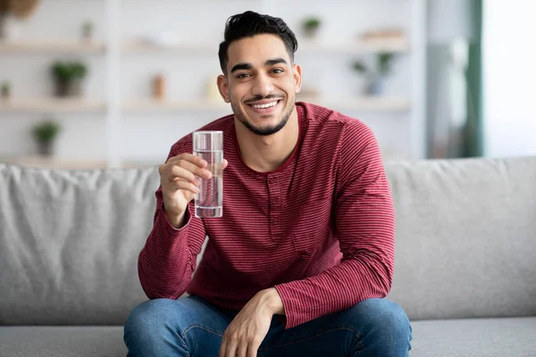 Cheerful arab guy sitting on couch, drinking water, copy space — Stock Photo, Image