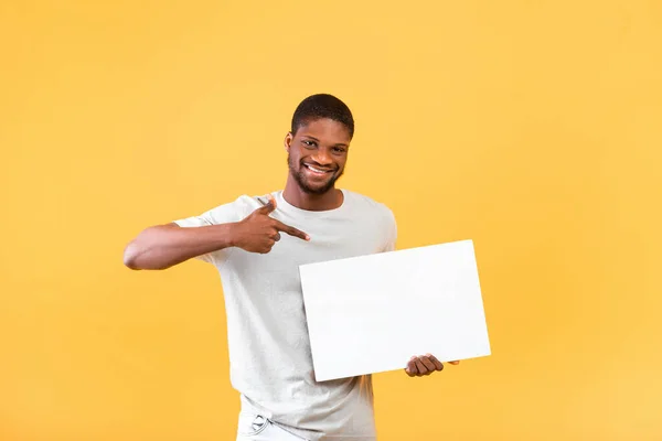 Werbetafel. Glücklicher schwarzer Kerl zeigt auf leeres weißes Poster mit Attrappe, steht über gelbem Hintergrund — Stockfoto