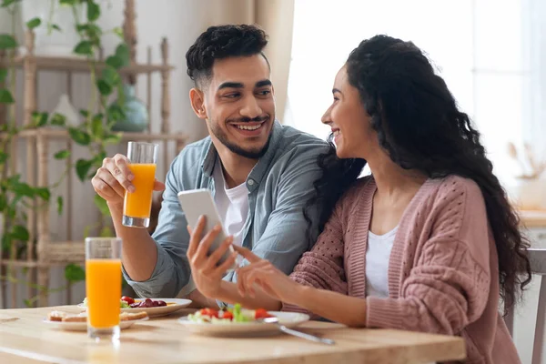 Glada arabiska par äta frukost tillsammans och använda smartphone i köket — Stockfoto