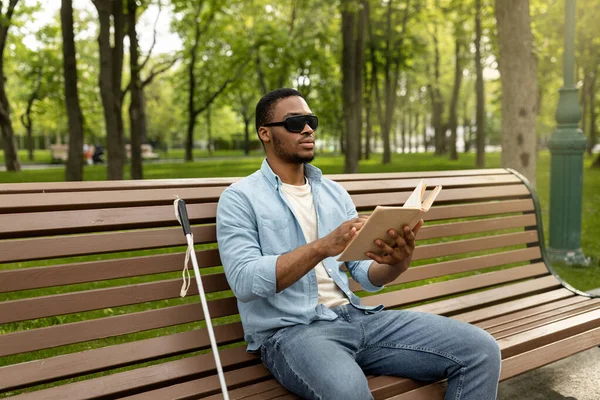 Joven hombre negro con discapacidad visual sentado en el banco en el parque de la ciudad, leyendo el libro Braille al aire libre — Foto de Stock