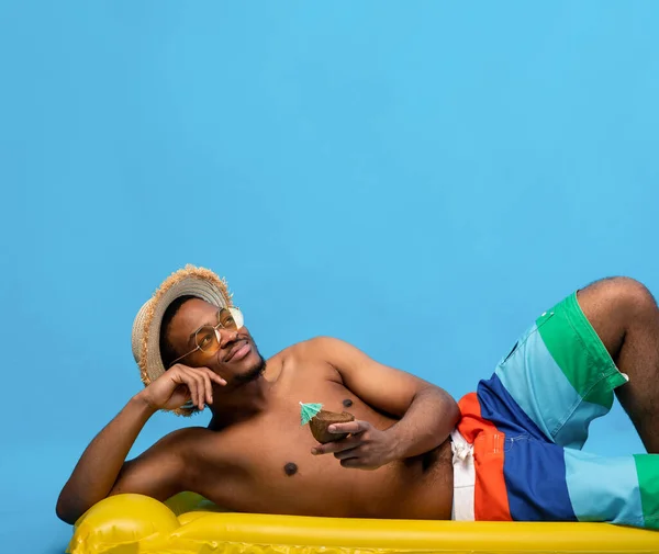 Hombre negro soñador tomando el sol en lilo inflable, bebiendo cóctel tropical de cáscara de coco sobre fondo azul —  Fotos de Stock