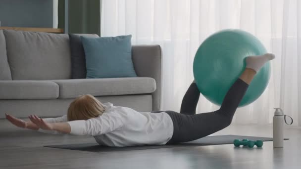 自宅で胃の上に寝そべってフィットボールを練習するシニア女性 — ストック動画