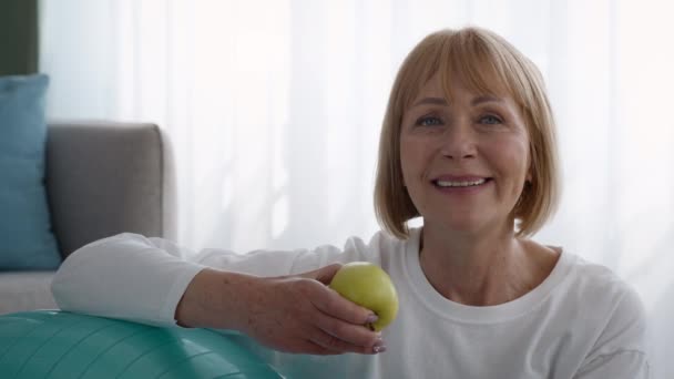 Senior Lady Holding Apple zitten in de buurt Fitball na training binnen — Stockvideo