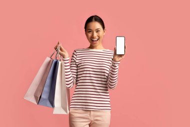 Heyecanlı Asyalı kadın elinde poşetlerle boş ekranla akıllı telefon gösterisi yapıyor.