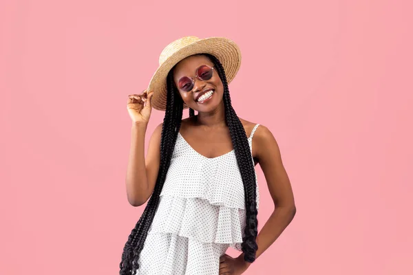 Pretty black lady with afro bunches wearing summer dress straw hat and sunglasses, smiling at camera on pink background — Stock Photo, Image