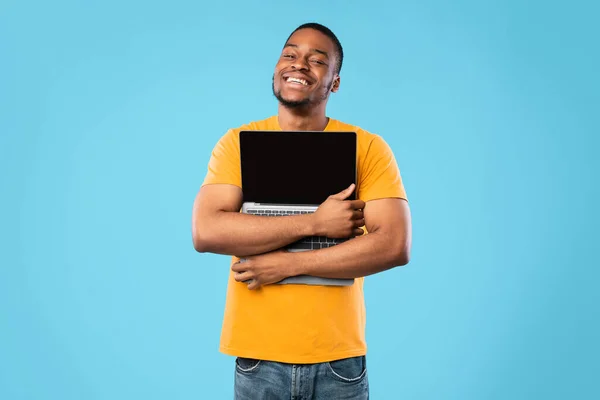 Hombre afroamericano abrazando el ordenador portátil con pantalla vacía, fondo azul —  Fotos de Stock
