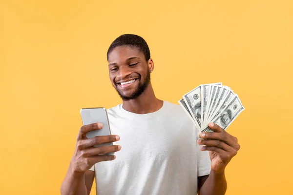 在网上赚钱和兑现的概念。快乐的黑人用智能手机，显示一堆钱，黄色背景 — 图库照片