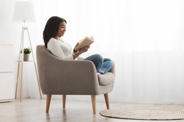 Helg-tidsfördriv. Avslappnad afrikansk amerikansk kvinna läser bok i bekväm fåtölj mot fönster, fritt utrymme — Stockfoto