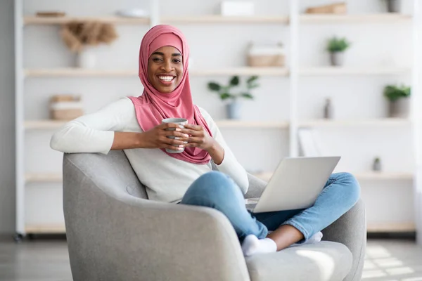 Ocio doméstico. Negro musulmana dama relajante en casa con ordenador portátil y café — Foto de Stock