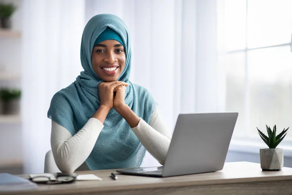 Happy Smiling Black Muslim Lady In Hijab Sitting At Desk With Laptop