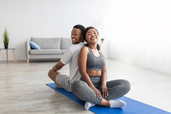 Gülümseyen Afrikalı Amerikalı çift yoga antrenmanından sonra dinleniyor, spor minderinde sırt sırta oturuyor, kameraya gülümsüyor. — Stok fotoğraf