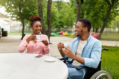 Tekerlekli sandalyedeki engelli genç adam açık kafede güzel bir kadınla buluşuyor, kahve içiyor, sohbetten zevk alıyor.
