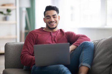 Gülümseyen Arap adam koltukta oturuyor ve dizüstü bilgisayar kullanıyor.