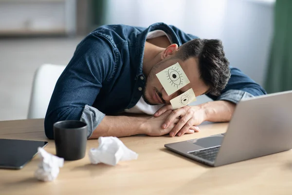 Exhausted arab guy with eyes drawing sleeping at workdesk — Stock Photo, Image