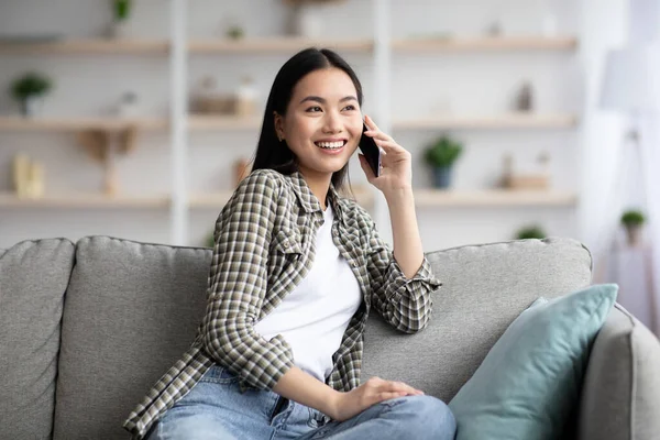 陽気なアジアの女性はソファに座ってスマートフォンで話して — ストック写真