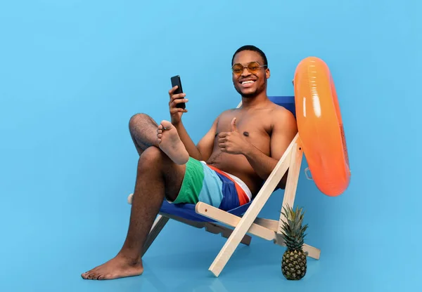 Homem negro alegre em roupa de banho sentado em cadeira de estar com celular, recomendando novo aplicativo móvel, mostrando o polegar para cima — Fotografia de Stock