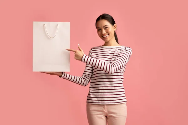 Place For Logo. Cheerful Korean Female Pointing At Blank Paper Shopping Bag