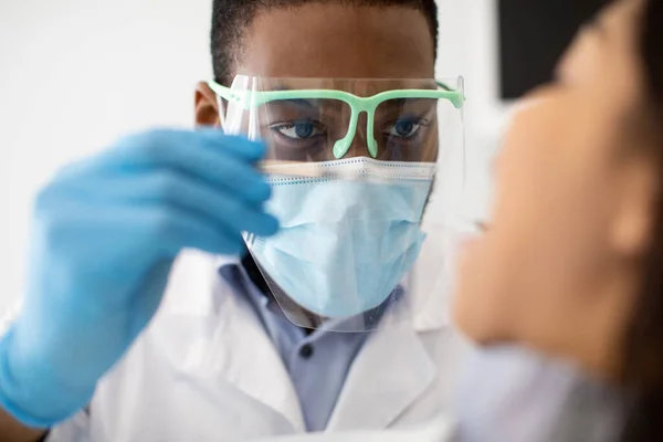 Dentista masculino preto na máscara protetora e no protetor facial examinando os dentes dos pacientes — Fotografia de Stock