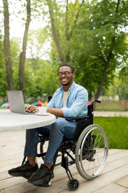 Şehir parkındaki kafede dizüstü bilgisayarla çalışan tekerlekli sandalyedeki neşeli, engelli siyahi adam internet üzerinden iş görüşmesi yapıyor.