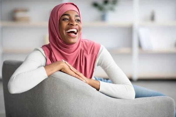 Vidám nevető fekete muszlim nő Hidzsábon ül a székben otthon — Stock Fotó