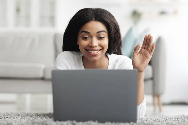 Felice donna nera fare videochiamate sul computer portatile a casa, agitando e sorridendo allo schermo mentre si trova sul pavimento in soggiorno — Foto Stock