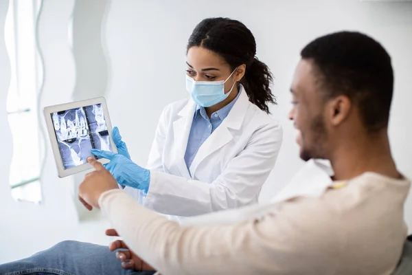 Μαύρη γυναίκα οδοντίατρος που δείχνει αρσενικό ασθενή δόντια ακτινογραφία εικόνα σε ψηφιακό δισκίο — Φωτογραφία Αρχείου