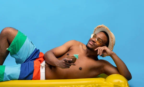 Chico negro con torso desnudo acostado sobre lilo inflable, disfrutando de agua de coco fresca o cóctel tropical, fondo azul —  Fotos de Stock