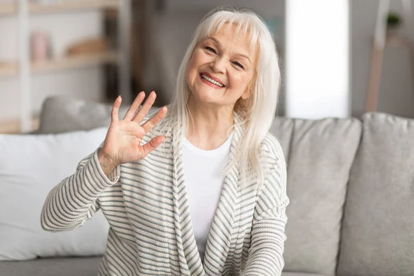 Зрелая женщина машет в камеру и улыбается, сидит на диване — стоковое фото